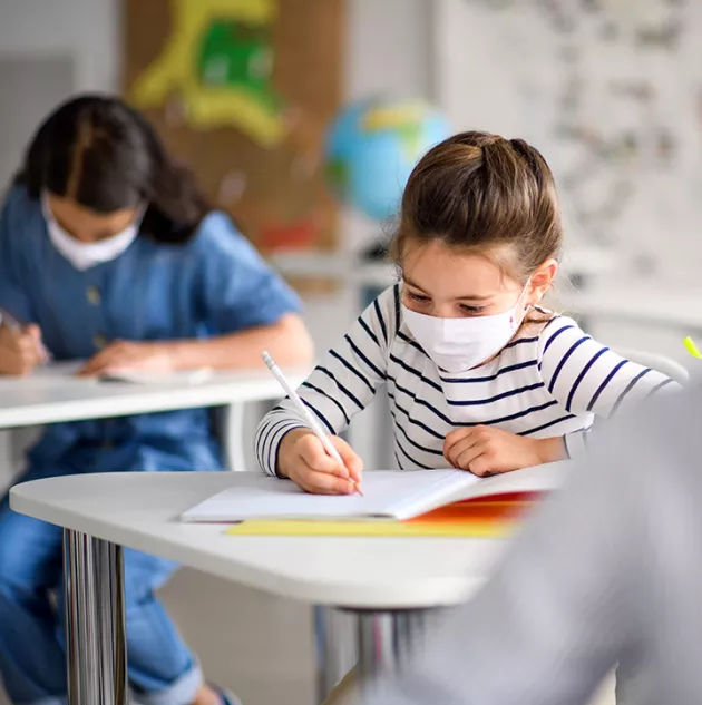 A girl writing at a school desk while wearing a mask.